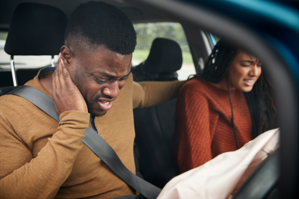 An injured man holds his neck in severe pain after a car accident while a woman looks distressed next to a deployed airbag, illustrating a whiplash or soft-tissue injury case for an Atlanta personal injury law firm.